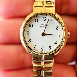 May include: A gold-toned Citizen Quartz watch with a white face and black numerals. The watch has a gold expanding band and a small crown. The watch face displays the Citizen Quartz logo and the text "JAPAN MOVT 5920-5010841 S".