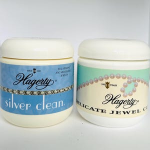 May include: Two Hagerty silver and jewelry cleaning jars. One jar is light blue with "silver clean" text. The other jar is cream-colored with "delicate jewel clean" text. Both jars have a white lid and the Hagerty logo.