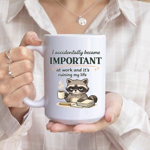 May include: White ceramic mug with the text "I accidentally became IMPORTANT at work and it's ruining my life." Features a cartoon raccoon with a coffee cup and stack of papers.