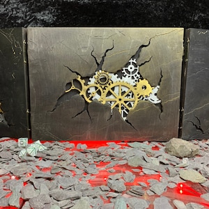 May include: A three-panel, black and gold, wooden box with a cracked surface revealing a set of gears. The box is sitting on a red surface covered in small, grey slate pieces. There are also four green dice in the foreground.