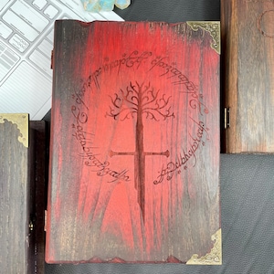 Könnte beinhalten: Ein hölzernes Buch mit rotem Einband, das ein Baum- und Schwertdesign mit Schriftzug zeigt. Die Ecken sind mit goldfarbenem Metall akzentuiert. Würfel und ein Spielblatt sind im Hintergrund zu sehen.