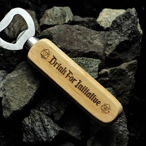 Puede incluir: Un abridor de botellas de madera con un abridor de tapas de metal. El mango est&aacute; grabado con el texto "Drink For Initiative" y un s&iacute;mbolo.