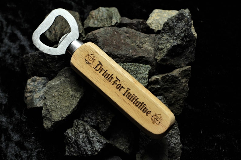 Puede incluir: Un abridor de botellas de madera con un abridor de tapas de metal. El mango est&aacute; grabado con el texto "Drink For Initiative" y un s&iacute;mbolo.