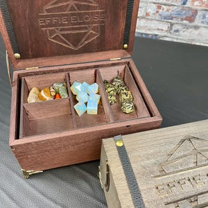 May include: Open wooden box with compartments, containing gemstones and dice. The box is dark brown with gold hardware and the text "EFFIE ELOISE" engraved on the lid. A second closed box is visible.