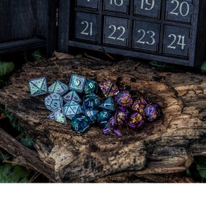 May include: A collection of multi-faceted polyhedral dice in various colours including silver, teal, and purple with gold flecks. The dice are arranged on a piece of dark, textured tree bark. A black wooden box with numbered slots 21 through 24 is visible.