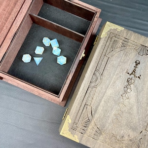 May include: A wooden dice box with a dark brown finish, featuring a compartment with a black interior and a set of iridescent dice. The box has a decorative design on the lid with brass corner accents.