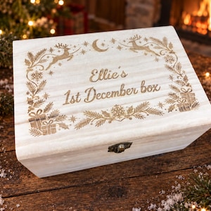 May include: A rectangular light-coloured wooden box with a hinged lid and a metal latch. The lid features laser-engraved holiday imagery including reindeer, pine branches, gifts, and a crescent moon, with the text Ellie's 1st December box.