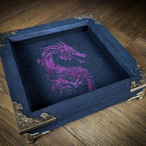 Peut inclure: Un plateau à dés carré en bois bleu foncé avec des coins de protection en laiton orné. L'intérieur présente un tissu noir avec un motif de dragon violet vif. Le plateau repose sur une surface en bois.