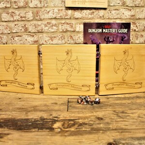 May include: A wooden Dungeon Master screen with five panels, each featuring a dragon design and the text "Dungeon Master". The screen is set up on a wooden table with dice scattered in front of it.