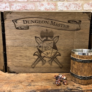Puede incluir: Una pantalla de Dungeon Master de madera con un diseño tallado de un dado de 20 caras, espadas cruzadas y un hacha. La pantalla tiene una pancarta que dice "Dungeon Master". También hay una taza de madera marrón y dados marrones en la imagen.