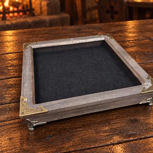 May include: A rectangular wooden tray with a weathered grey finish, featuring ornate brass-toned metal corner protectors and decorative claw-style feet. The interior base of the tray is lined with a solid black felt material.