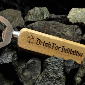 Puede incluir: Un abridor de botellas de madera con un abridor de metal y un mango grabado con "Drink For Initiative" y un s&iacute;mbolo D20.