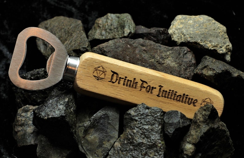 Puede incluir: Un abridor de botellas de madera con un abridor de metal y un mango grabado con "Drink For Initiative" y un s&iacute;mbolo D20.