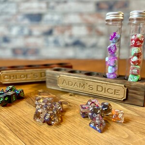 May include: A wooden dice tower with two glass tubes filled with colorful dice. The tower has a sign that reads "Adam's Dice". There is a second wooden dice tower in the background with a sign that reads "Chloe's D".