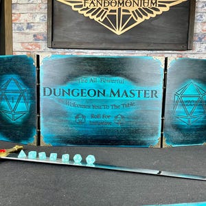 Pode incluir: Um ecrã de Mestre de Masmorras de madeira com acabamento em verde-azulado e preto, com o texto "Dungeon Master" e desenhos geométricos. Inclui uma espada e um conjunto de dados. O ecrã tem cerca de 76 cm de largura.