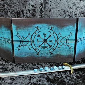 Könnte beinhalten: Ein dreiteiliger Tischspielbildschirm mit türkis-schwarzem Finish und einem schwarzen geometrischen Design. Ein Silberschwert mit goldenem Griff und ein Satz blauer Würfel im Vordergrund deuten auf ein Fantasy-Rollenspiel-Thema hin.