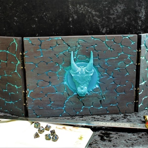May include: A three-panel wooden game board with a blue dragon head sculpted into the centre panel. The panels are painted black with a cracked blue and green effect. The board is set up on a black surface with a sword and dice in front of it.