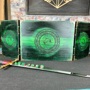 May include: A three-panel wooden game board with a green stained finish. The centre panel features a circular design with the text "The Great Dice Hall of the Young, North, East, South, West, and the Rolling of the Dice."  The board is surrounded by a gold trim.  A sword and several green dice are in the foreground.