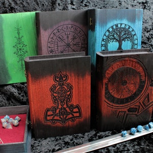 May include: Four wooden boxes with different designs. The boxes are all black with a different coloured stain on the front. The designs are all Norse-inspired and include a tree of life, a compass, a hammer, and a circle with runes. The boxes are all closed and sitting on a black surface.
