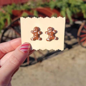 May include: A pair of brown gingerbread man earrings with white icing details. The earrings are on a cream-colored card. The background is blurred, with green foliage and a red wheel visible.