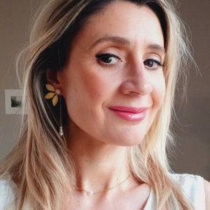 Op de afbeelding: Een vrouw draagt een wit topje met een delicate gouden ketting met een enkele parel hanger. Ze draagt gouden oorbellen met een bloemmotief en hangende parels.