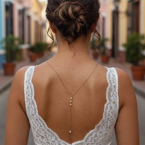Può includere: Vista posteriore di una donna che indossa un abito di pizzo bianco con schiena scoperta. Una delicata collana d'oro con tre perle pende lungo la schiena. I capelli della donna sono raccolti in uno chignon. Lo sfondo è una strada colorata.