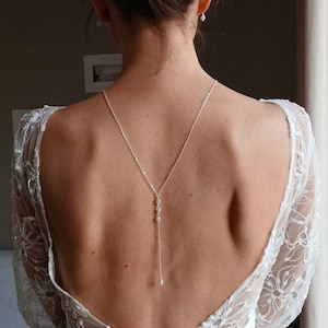 Puede incluir: Un collar de espalda plateado con un diseño delicado, con pequeños adornos brillantes. El collar cuelga por la espalda, complementando un vestido blanco con mangas de encaje. La imagen muestra la espalda de una persona que lleva el collar.