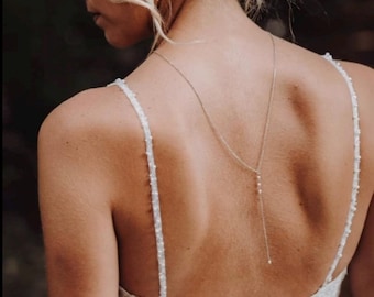 Könnte beinhalten: Eine Braut in einem weißen Spitzen-Hochzeitskleid mit tiefem Rücken und einer zarten Perlenkette. Das Kleid hat ein figurbetontes Oberteil und einen fließenden Rock. Die Haare der Braut sind zu einem niedrigen Dutt mit einem Blumenhaarband gesteckt.