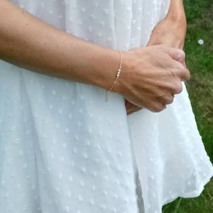 Puede incluir: Un delicado brazalete de cadena dorada con pequeñas cuentas blancas. El brazalete se lleva en la muñeca.
