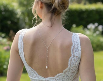 Può includere: Una collana d'argento con un pendente a perla scende lungo la schiena di un abito di pizzo bianco. L'abito ha la schiena scoperta e delicati dettagli in pizzo. Lo sfondo è un giardino verde sfocato.