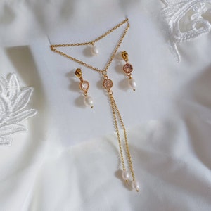 Puede incluir: Un collar de oro con una cadena delicada y dos pequeñas perlas. El collar se combina con pendientes a juego que presentan una pequeña perla y una piedra de cristal transparente.