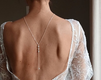 Può includere: Una donna indossa un abito bianco con la schiena in pizzo e una delicata collana di schiena argentata con piccoli accenti di cristallo.