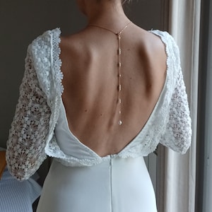 Peut inclure: Une robe de mariée en dentelle blanche avec un dos nu et un délicat collier de dos en perles et chaîne dorée.