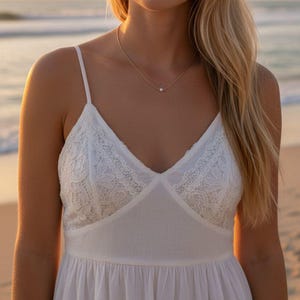 Op de afbeelding: Een witte zomerjurk met kanten details op het lijfje en spaghettibandjes. Een delicate gouden ketting met een enkele parel hanger wordt gedragen. De achtergrond toont een strandscène met de oceaan en een zonsondergang.