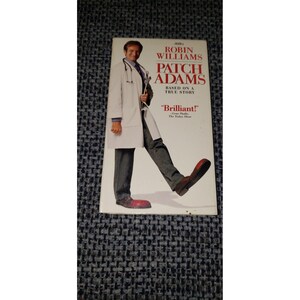 May include: A VHS tape cover for the movie Patch Adams featuring Robin Williams in a white lab coat and stethoscope, wearing oversized red clown shoes. The text reads Robin Williams, Patch Adams, Based on a True Story, and Brilliant! Gene Shalit.