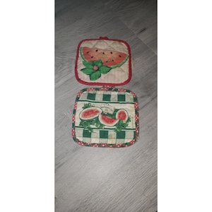 May include: Two square quilted pot holders featuring watermelon designs. The top holder has a single watermelon slice on a striped background. The bottom holder displays watermelon slices and green leaves on a green and white checkered pattern.