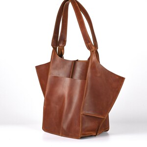 Puede incluir: Un bolso tote de cuero marr&oacute;n con dos asas. El bolso tiene un compartimento grande y abierto y un bolsillo exterior. El cuero tiene una veta natural y un color marr&oacute;n rojizo c&aacute;lido. El bolso est&aacute; dise&ntilde;ado con una est&eacute;tica moderna y minimalista.