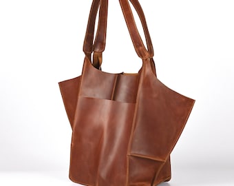 Può includere: Una borsa tote in pelle marrone con due manici. La borsa ha un ampio scomparto aperto e una tasca esterna. La pelle ha una grana naturale e un colore marrone-rossastro caldo. La borsa è progettata con un'estetica moderna e minimalista.