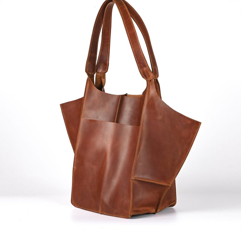 Puede incluir: Un bolso tote de cuero marr&oacute;n con dos asas. El bolso tiene un compartimento grande y abierto y un bolsillo exterior. El cuero tiene una veta natural y un color marr&oacute;n rojizo c&aacute;lido. El bolso est&aacute; dise&ntilde;ado con una est&eacute;tica moderna y minimalista.