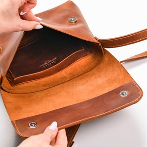 May include: A tan leather crossbody bag with a fold-over flap, featuring two silver-toned magnetic snap closures and a long shoulder strap. The interior reveals a dark brown leather pocket embossed with a logo on the back wall of the main compartment.