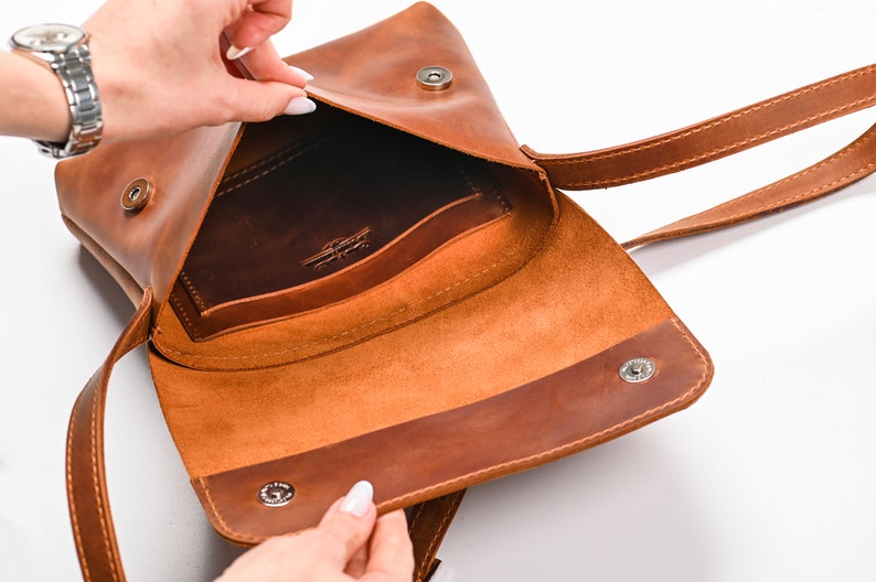 May include: A tan leather crossbody bag with a fold-over flap, featuring two silver-toned magnetic snap closures and a long shoulder strap. The interior reveals a dark brown leather pocket embossed with a logo on the back wall of the main compartment.