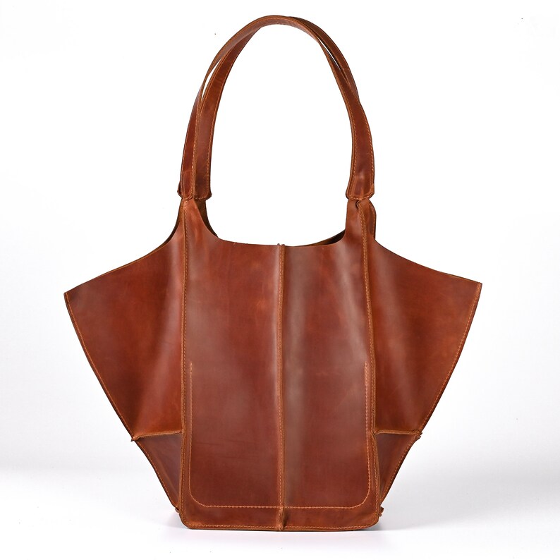 Pode incluir: Uma bolsa tote de couro marrom com uma parte superior larga e aberta e duas al&ccedil;as longas. A bolsa tem um formato angular &uacute;nico e um design simples e minimalista.