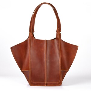 Puede incluir: Un bolso tote de cuero marr&oacute;n con dos asas y un compartimento abierto grande. El bolso tiene forma rectangular con un bolsillo en la parte delantera. El cuero tiene un aspecto envejecido.