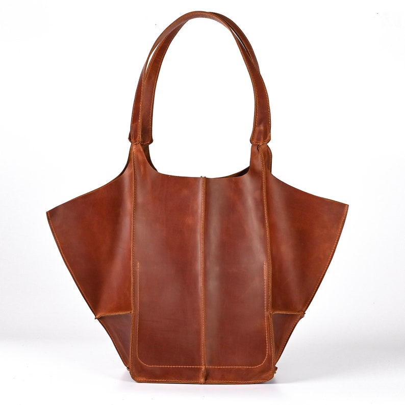 Puede incluir: Un bolso tote de cuero marr&oacute;n con dos asas y un compartimento abierto grande. El bolso tiene forma rectangular con un bolsillo en la parte delantera. El cuero tiene un aspecto envejecido.