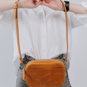 May include: A tan leather crossbody bag with a zipper closure and a small pocket on the front. The bag has a brown leather strap and a silver buckle. The bag is worn by a person wearing a white shirt and blue jeans.