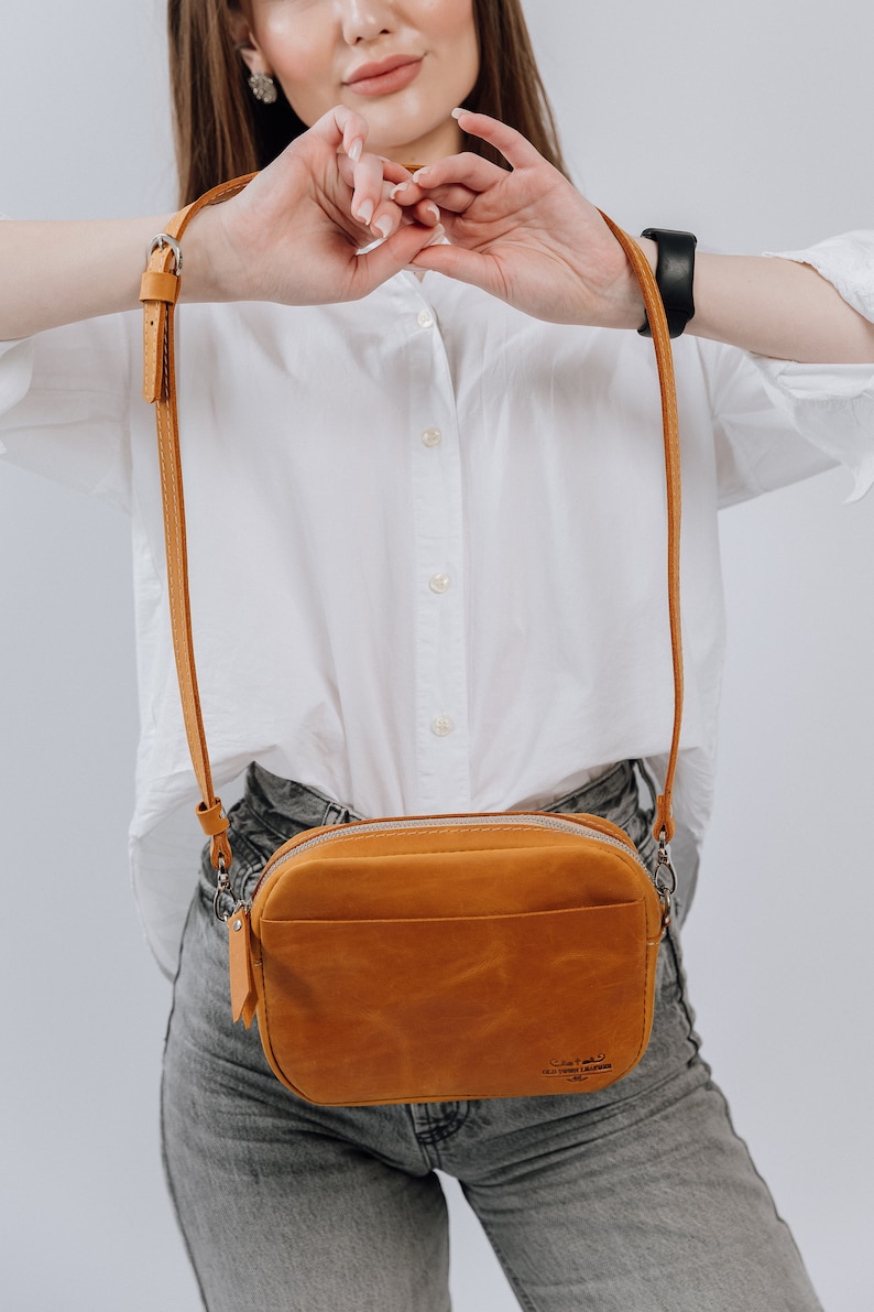 May include: A tan leather crossbody bag with a zipper closure and a small pocket on the front. The bag has a brown leather strap and a silver buckle. The bag is worn by a person wearing a white shirt and blue jeans.