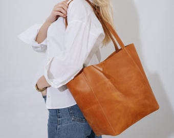May include: A large, rectangular, tan leather tote bag with long handles. The bag has visible stitching and a simple, classic design. The bag is carried over the shoulder.