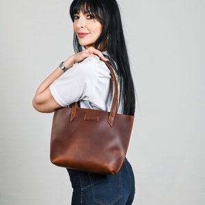 May include: A brown leather tote bag with a simple design. The bag has a single compartment and two short handles.