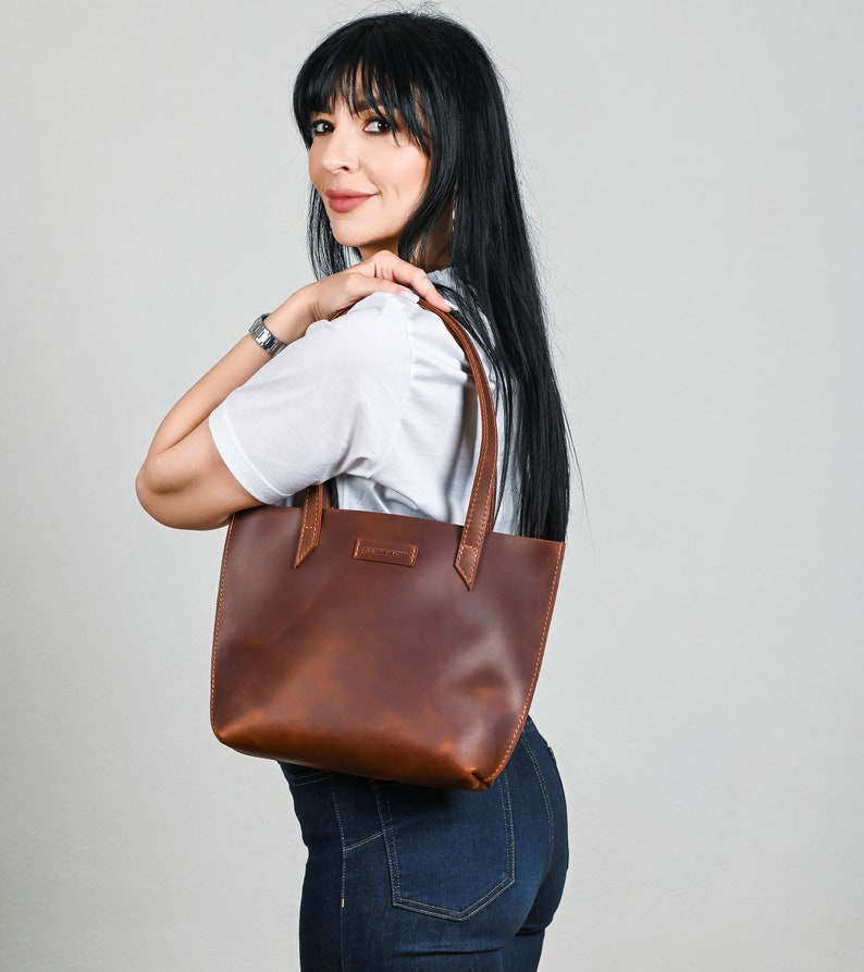 May include: A brown leather tote bag with a simple design. The bag has a single compartment and two short handles.