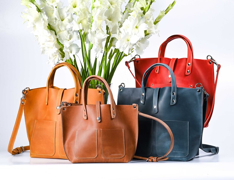 May include: Four leather tote bags in different colors and sizes. The bags have a top handle and a shoulder strap. The bags are arranged in a row on a white surface.
